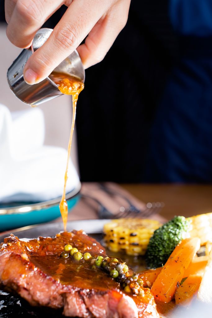 Close-up of steak with peppercorn sauce being poured, accompanied by vegetables, showcasing gourmet dining.