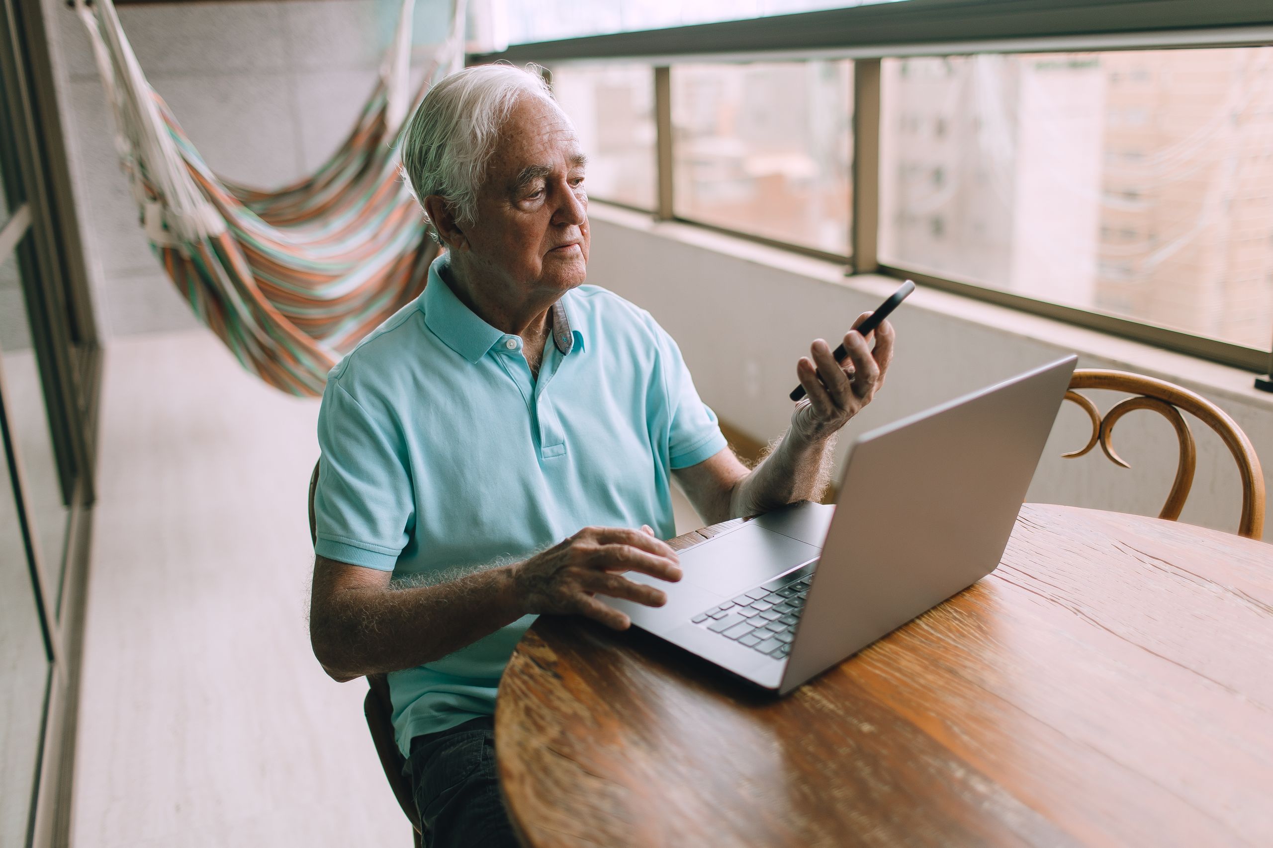 Elderly man multitasking with laptop and smartphone indoors, showcasing modern technology use.