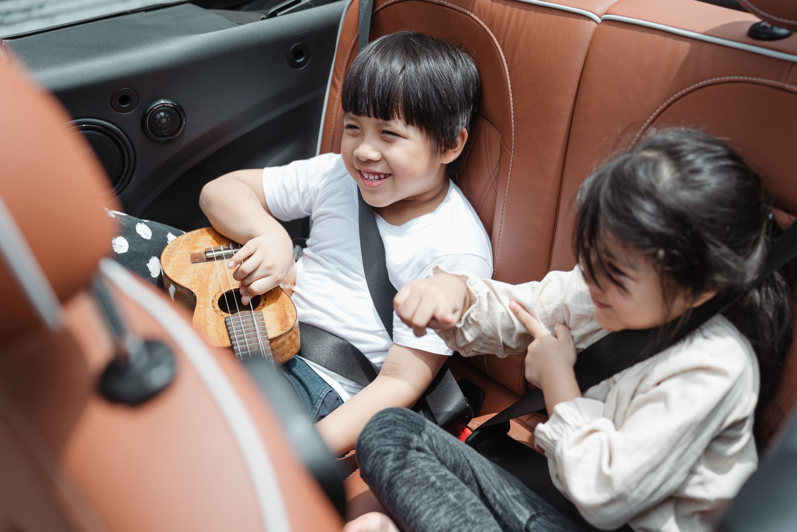 From above smiling ethnic boy and girl in casual outfits sitting fastened in passenger seats with ukulele during road trip together