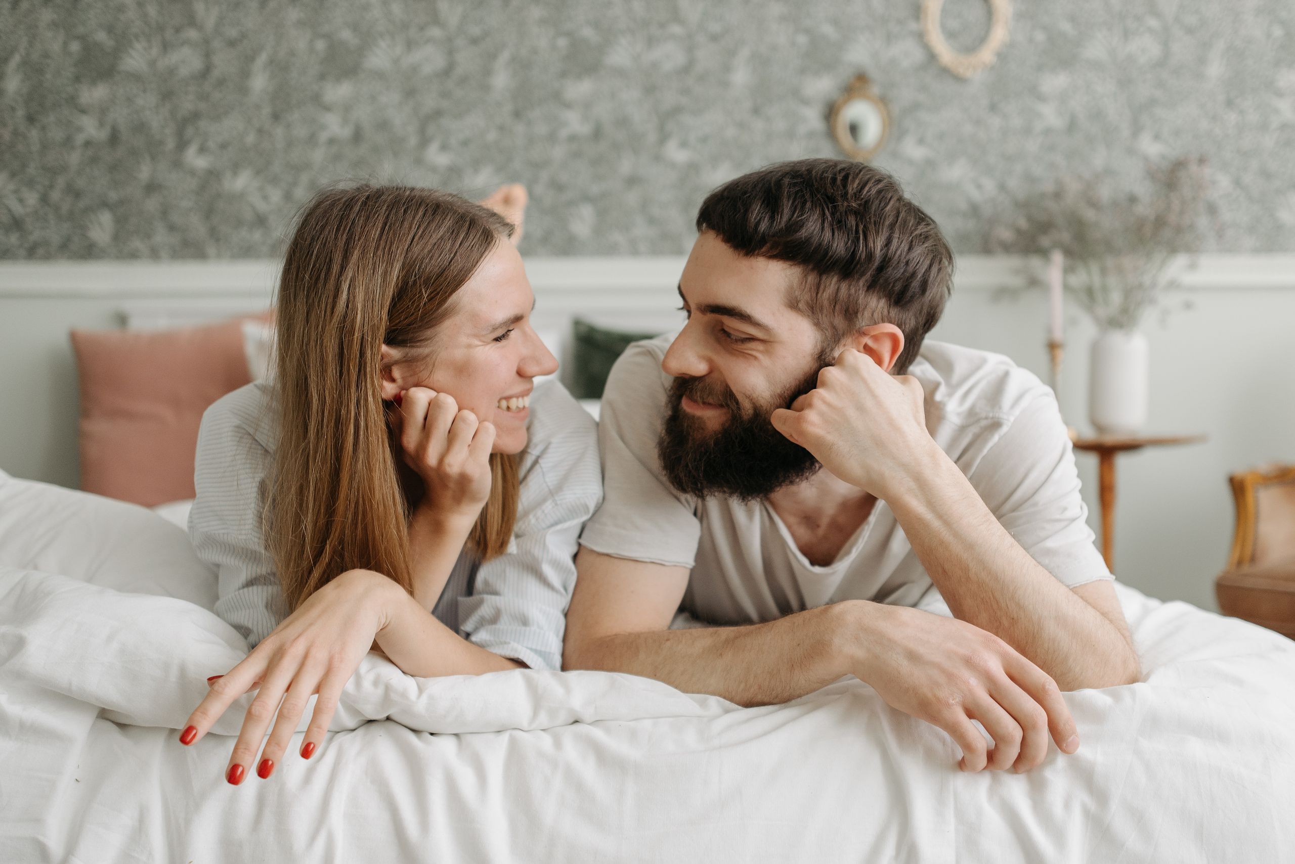 Happy couple lying face to face on a bed, sharing smiles in a cozy and intimate bedroom setting.
