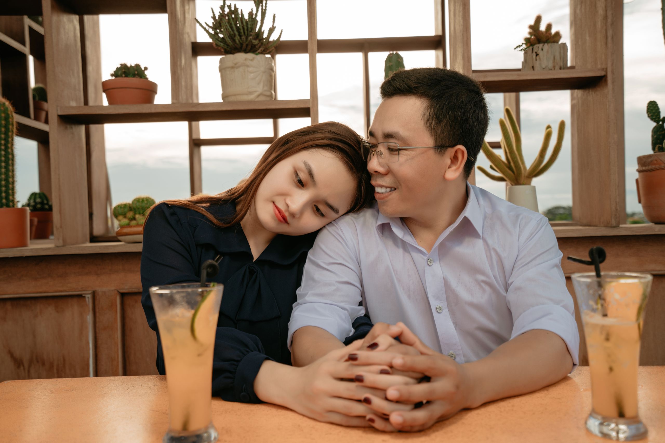 Positive ethnic couple in elegant outfits sitting at table with cocktails on date in cafe and holding hands while hugging in daylight with potted plants on shelves on background