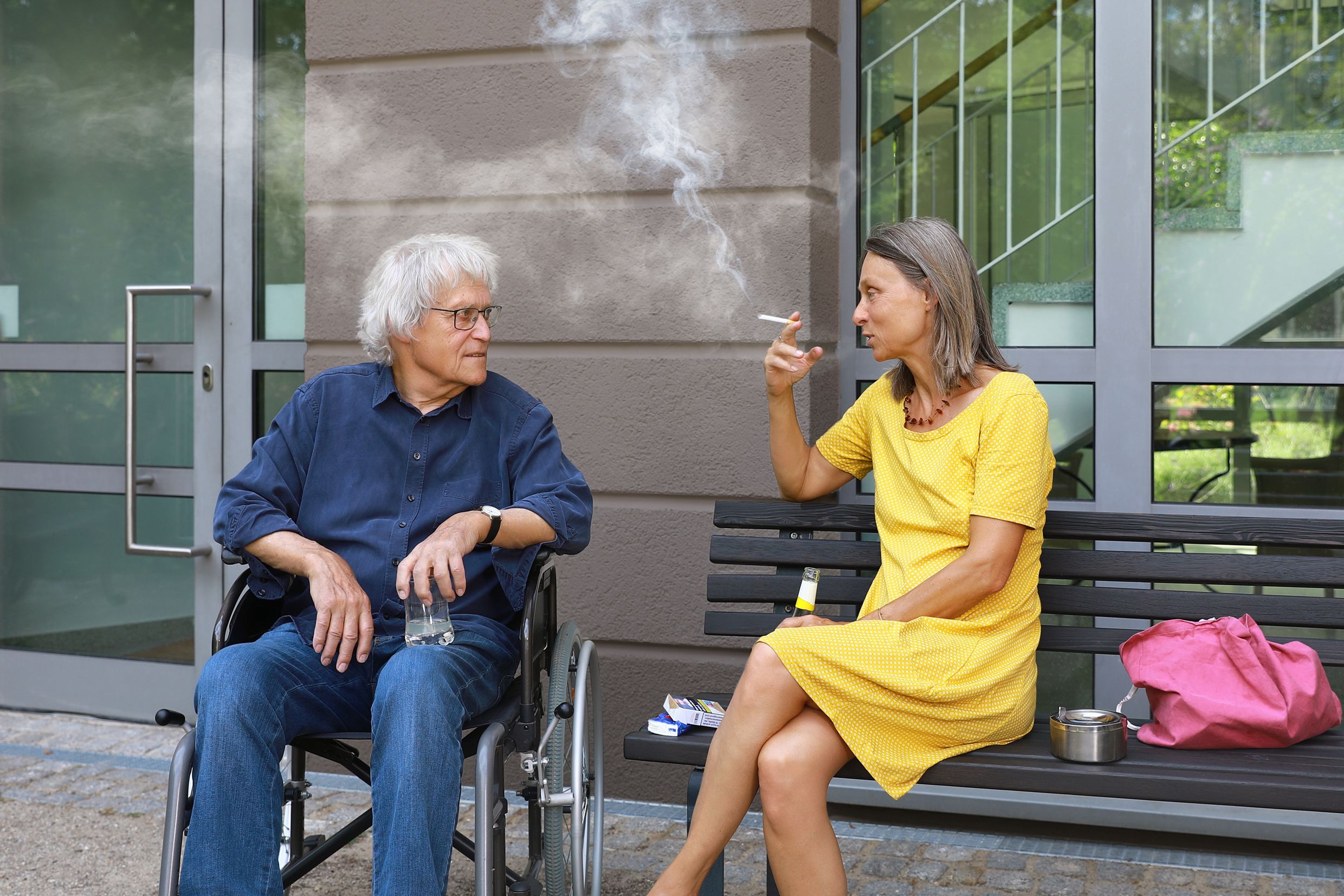 Senior man in wheelchair and woman in yellow dress smoking outside urban building.