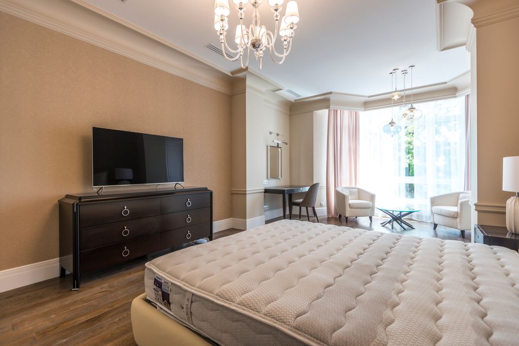 Spacious modern bedroom with chandelier and flat screen TV, featuring a minimalist and elegant design.