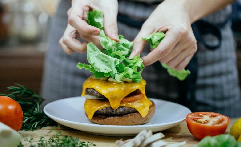 Unrecognizable female cook putting fresh lettuce on grilled cheese while preparing burger with cutlets at table with tomatoes against blurred background