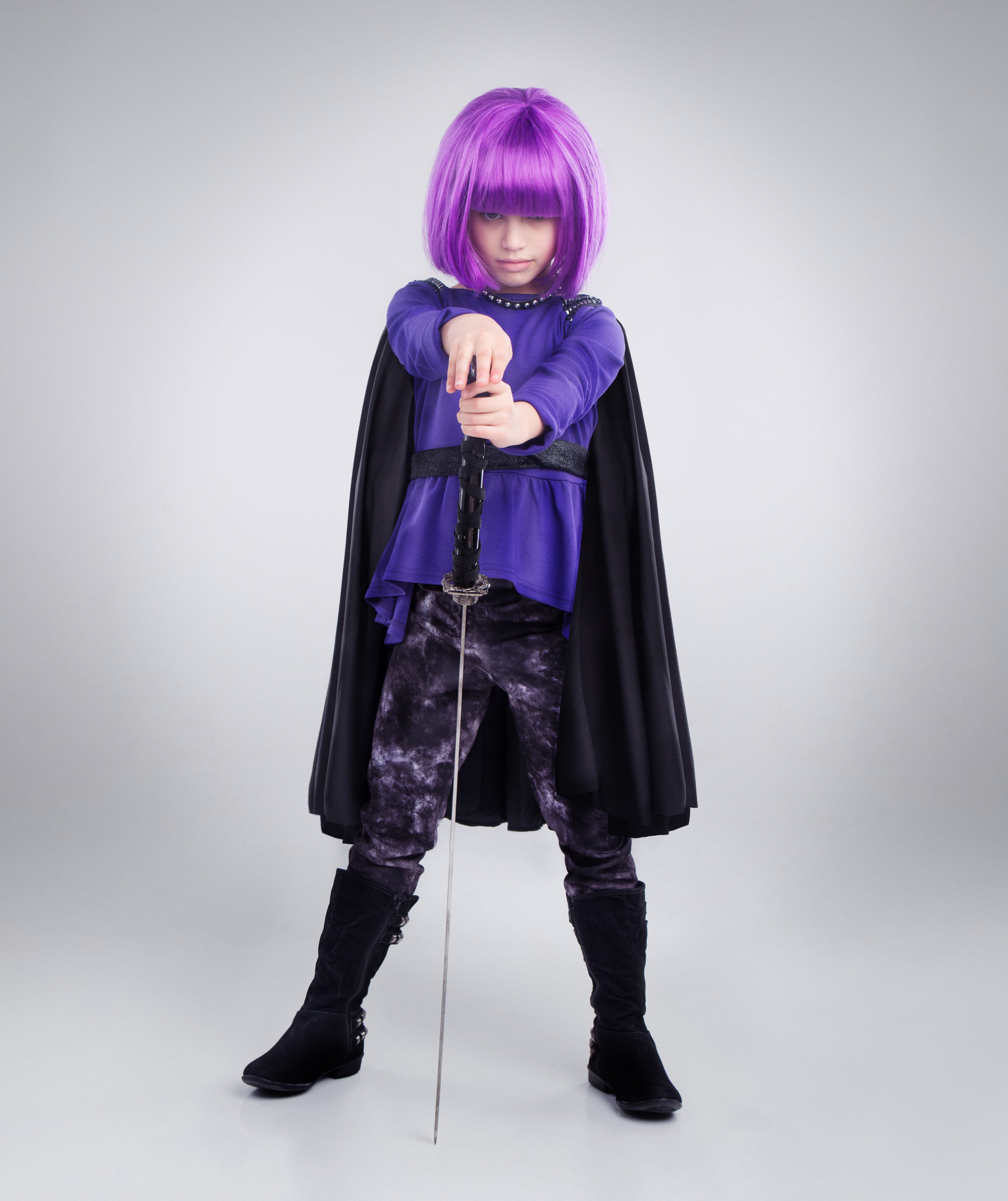 Little girl, superhero and halloween dress up posing with sword against a grey studio background. Portrait of girl child playing super hero with purple hair, costume or cosplay of a warrior vigilante